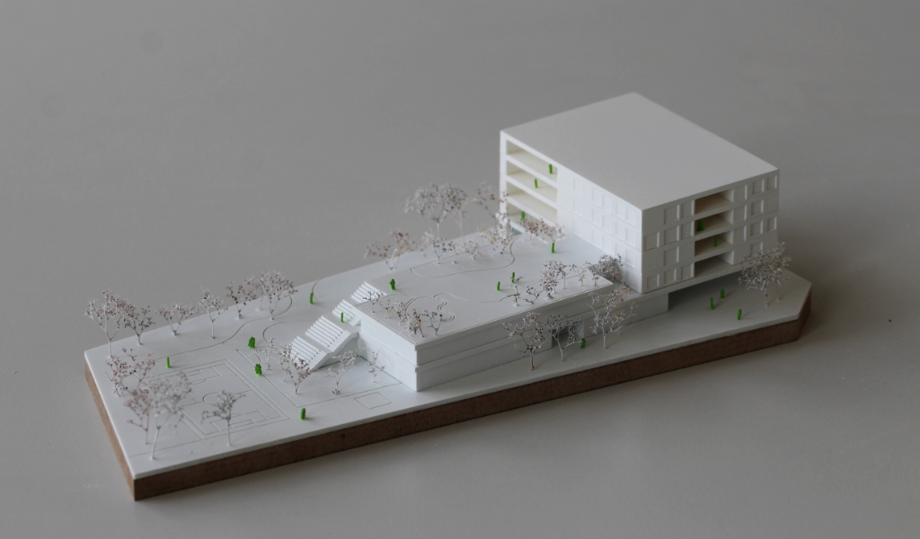 Weißes Wettbewerbsmodell eines Schulbaus. Längliche weiße dünne Grundplatte mit eingravierten Freianlagen und weiß lackierten Bäumen aus getrockneten Pflanzen. Auf einem langgestreckten zweigeschossigen Grundkörper, dessen Dach über eine große Freitreppe begehbar ist und worauf auch Bäume gepflanzt sind, sitzt etwas versetzt und um 90 Grad gedreht ein viergeschossiger Baukörper mit übereinander liegenden Loggien.