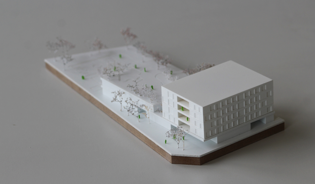 Weißes Wettbewerbsmodell eines Schulbaus. Längliche weiße dünne Grundplatte mit eingravierten Freianlagen und weiß lackierten Bäumen aus getrockneten Pflanzen. Auf einem langgestreckten zweigeschossigen Grundkörper, dessen Dach über eine große Freitreppe begehbar ist und worauf auch Bäume gepflanzt sind, sitzt etwas versetzt und um 90 Grad gedreht ein viergeschossiger Baukörper mit übereinander liegenden Loggien.