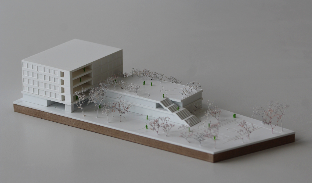Weißes Wettbewerbsmodell eines Schulbaus. Längliche weiße dünne Grundplatte mit eingravierten Freianlagen und weiß lackierten Bäumen aus getrockneten Pflanzen. Auf einem langgestreckten zweigeschossigen Grundkörper, dessen Dach über eine große Freitreppe begehbar ist und worauf auch Bäume gepflanzt sind, sitzt etwas versetzt und um 90 Grad gedreht ein viergeschossiger Baukörper mit übereinander liegenden Loggien.