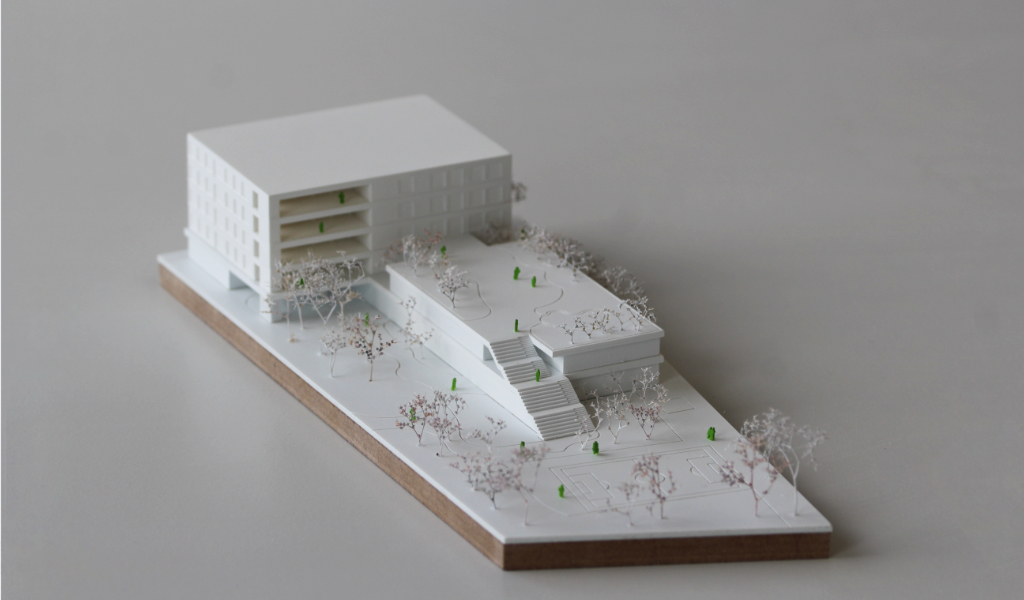 Weißes Wettbewerbsmodell eines Schulbaus. Längliche weiße dünne Grundplatte mit eingravierten Freianlagen und weiß lackierten Bäumen aus getrockneten Pflanzen. Auf einem langgestreckten zweigeschossigen Grundkörper, dessen Dach über eine große Freitreppe begehbar ist und worauf auch Bäume gepflanzt sind, sitzt etwas versetzt und um 90 Grad gedreht ein viergeschossiger Baukörper mit übereinander liegenden Loggien.