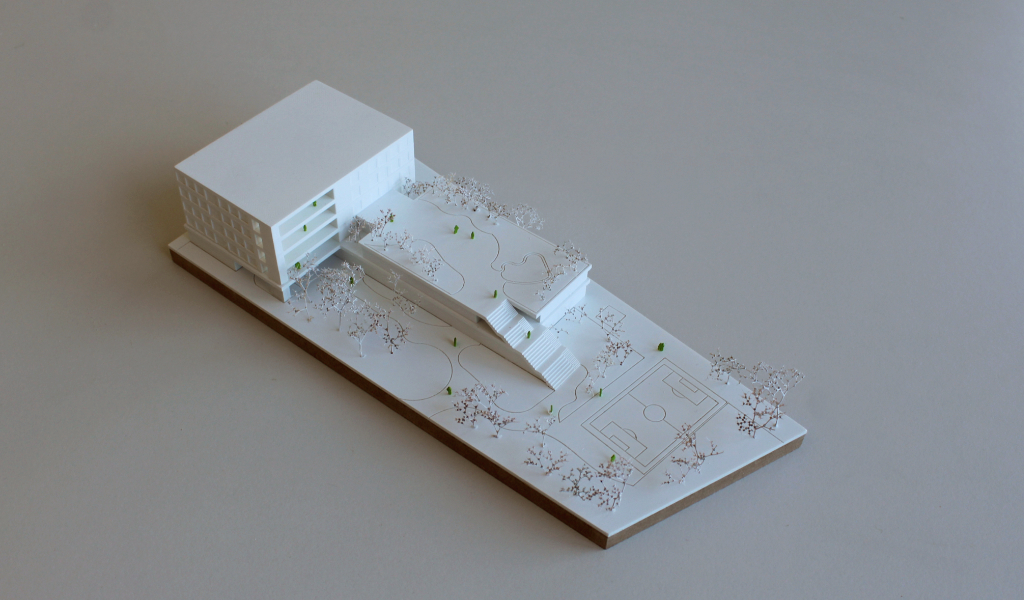 Weißes Wettbewerbsmodell eines Schulbaus. Längliche weiße dünne Grundplatte mit eingravierten Freianlagen und weiß lackierten Bäumen aus getrockneten Pflanzen. Auf einem langgestreckten zweigeschossigen Grundkörper, dessen Dach über eine große Freitreppe begehbar ist und worauf auch Bäume gepflanzt sind, sitzt etwas versetzt und um 90 Grad gedreht ein viergeschossiger Baukörper mit übereinander liegenden Loggien.