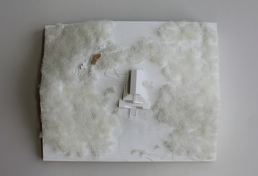 weißes Architekturmodell von der Walhalla und Umgebung. Der Höhenverlauf ist mittels Höhenschichten dargestellt, zur Dargestellung der Bäume ist weißer Pflanzenschaum flächig verklebt. Der Neubau des Besucherzentrums liegt nordöstlich von der Walhalla und besteht aus einem weißen Sockel auf dem ein kleines Holzvolumen mit mehreren Dachverschneidungen sitzt. Draufsicht von oben.