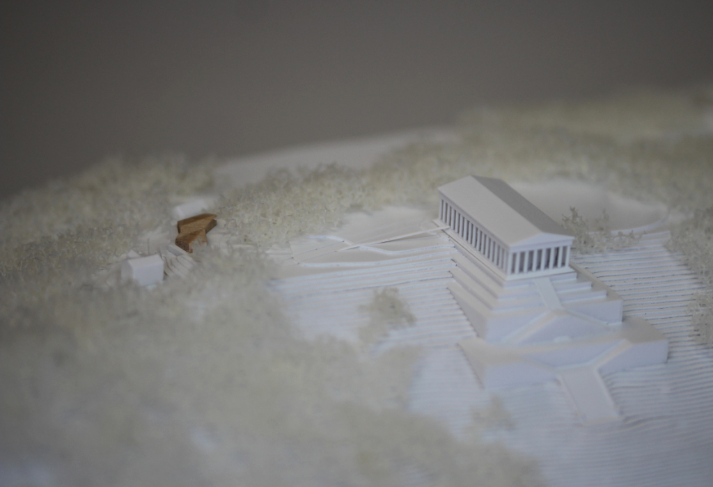 weißes Architekturmodell von der Walhalla und Umgebung. Der Höhenverlauf ist mittels Höhenschichten dargestellt, zur Dargestellung der Bäume ist weißer Pflanzenschaum flächig verklebt. Der Neubau des Besucherzentrums liegt nordöstlich von der Walhalla und besteht aus einem weißen Sockel auf dem ein kleines Holzvolumen mit mehreren Dachverschneidungen sitzt. Draufsicht von Südosten. Nahaufnahme