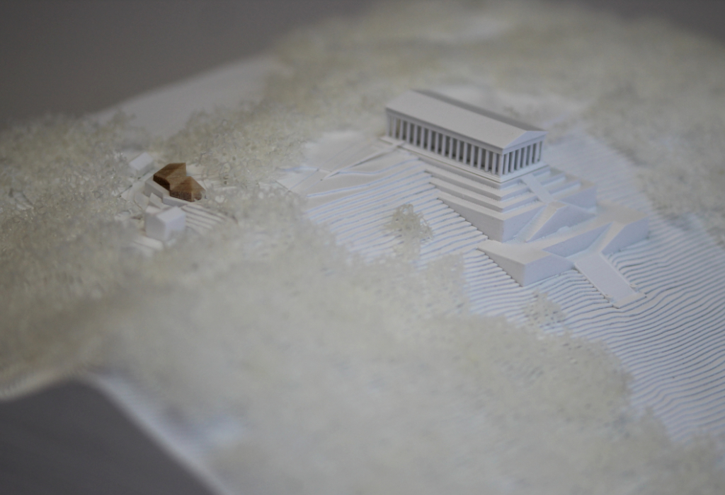 weißes Architekturmodell von der Walhalla und Umgebung. Der Höhenverlauf ist mittels Höhenschichten dargestellt, zur Dargestellung der Bäume ist weißer Pflanzenschaum flächig verklebt. Der Neubau des Besucherzentrums liegt nordöstlich von der Walhalla und besteht aus einem weißen Sockel auf dem ein kleines Holzvolumen mit mehreren Dachverschneidungen sitzt. Draufsicht von Südosten. Nahaufnahme.