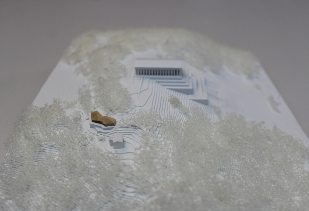 weißes Architekturmodell von der Walhalla und Umgebung. Der Höhenverlauf ist mittels Höhenschichten dargestellt, zur Dargestellung der Bäume ist weißer Pflanzenschaum flächig verklebt. Der Neubau des Besucherzentrums liegt nordöstlich von der Walhalla und besteht aus einem weißen Sockel auf dem ein kleines Holzvolumen mit mehreren Dachverschneidungen sitzt. Draufsicht von Osten.