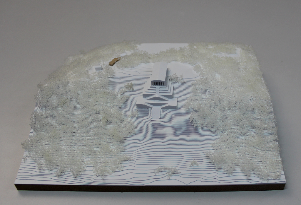 weißes Architekturmodell von der Walhalla und Umgebung. Der Höhenverlauf ist mittels Höhenschichten dargestellt, zur Dargestellung der Bäume ist weißer Pflanzenschaum flächig verklebt. Der Neubau des Besucherzentrums liegt nordöstlich von der Walhalla und besteht aus einem weißen Sockel auf dem ein kleines Holzvolumen mit mehreren Dachverschneidungen sitzt. Draufsicht von Süden.