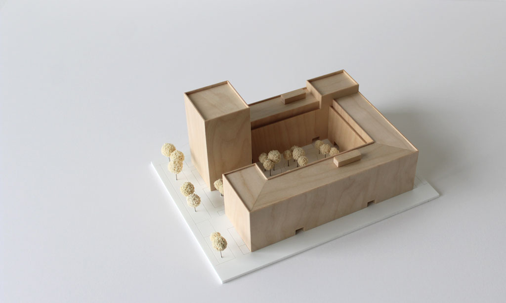 Volumenmodell aus Holz auf einer weißen Grundplatte mit Bäumen aus Schwammgummi. Blockrandbebauung mit Innenhof und einem höheren Turm an einer Ecke.