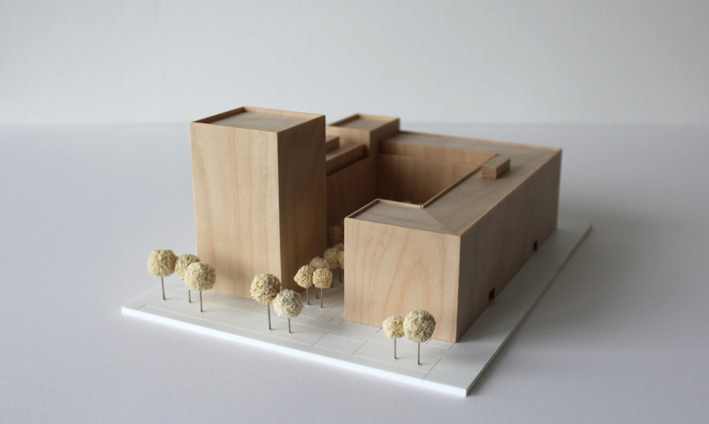 Volumenmodell aus Holz auf einer weißen Grundplatte mit Bäumen aus Schwammgummi. Blockrandbebauung mit Innenhof und einem höheren Turm an einer Ecke.