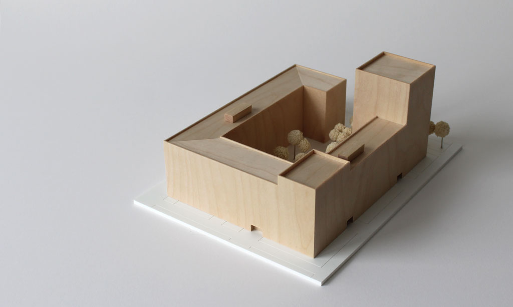 Volumenmodell aus Holz auf einer weißen Grundplatte mit Bäumen aus Schwammgummi. Blockrandbebauung mit Innenhof und einem höheren Turm an einer Ecke.