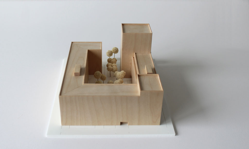 Volumenmodell aus Holz auf einer weißen Grundplatte mit Bäumen aus Schwammgummi. Blockrandbebauung mit Innenhof und einem höheren Turm an einer Ecke.