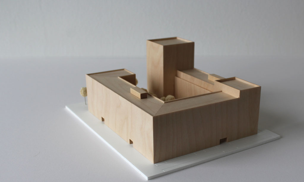 Volumenmodell aus Holz auf einer weißen Grundplatte mit Bäumen aus Schwammgummi. Blockrandbebauung mit Innenhof und einem höheren Turm an einer Ecke.