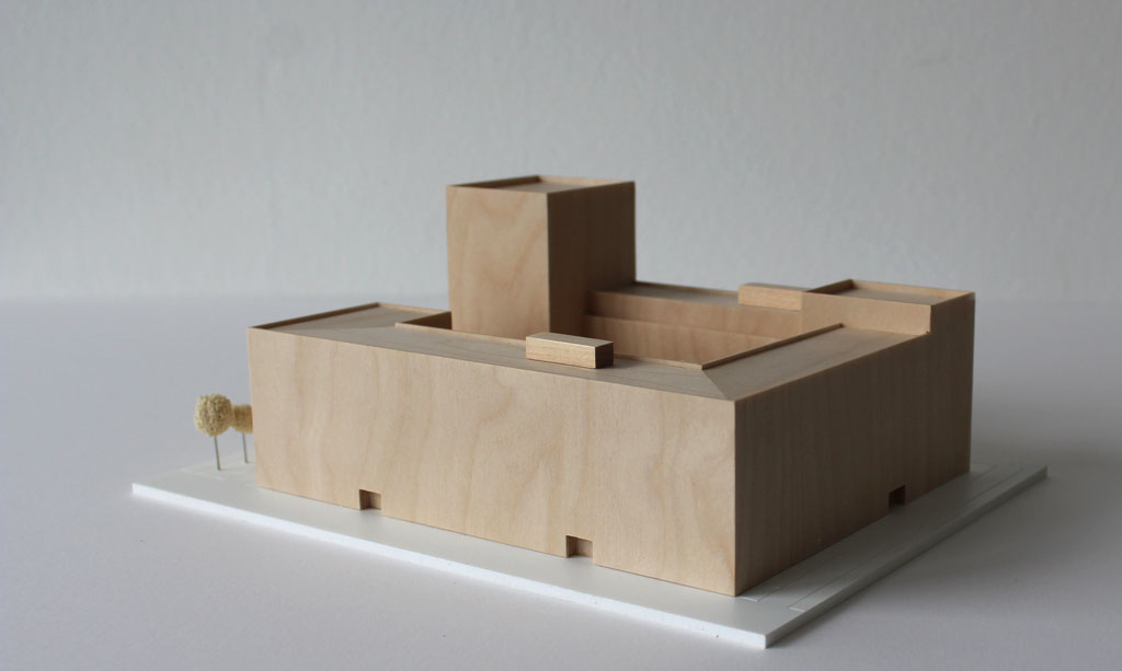 Volumenmodell aus Holz auf einer weißen Grundplatte mit Bäumen aus Schwammgummi. Blockrandbebauung mit Innenhof und einem höheren Turm an einer Ecke.