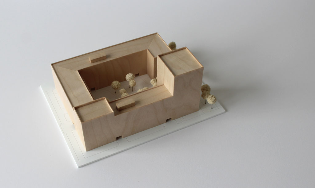 Volumenmodell aus Holz auf einer weißen Grundplatte mit Bäumen aus Schwammgummi. Blockrandbebauung mit Innenhof und einem höheren Turm an einer Ecke.