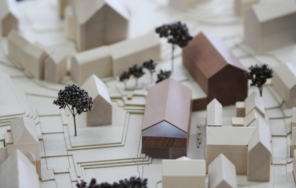Modell für Architektenwettbewerb mit Höhenschichten aus gelblicher Finnpappe und Gebäuden aus Lindenholz. Der Neubau des Rathauses besteht aus zwei Gebäuden mit Satteldach und ist aus rötlichem Elsbeerenholz und mattiertem Acyl. Die Bäume sind aus dunkelbraunem getrocknetem Planzenmaterial.