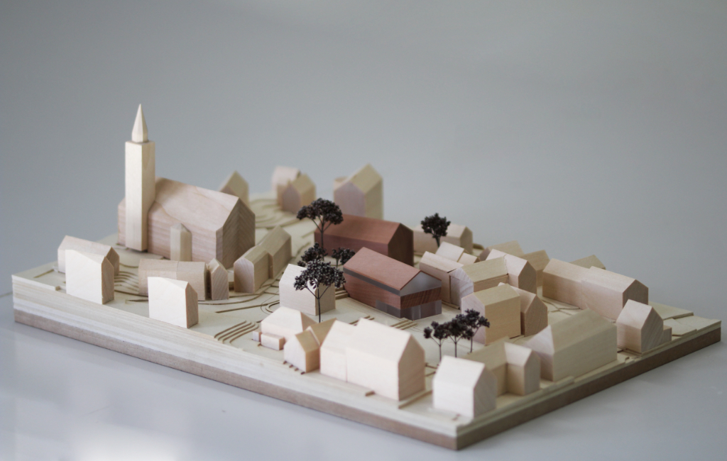 Modell für Architektenwettbewerb mit Höhenschichten aus gelblicher Finnpappe und Gebäuden aus Lindenholz. Der Neubau des Rathauses besteht aus zwei Gebäuden mit Satteldach und ist aus rötlichem Elsbeerenholz und mattiertem Acyl. Die Bäume sind aus dunkelbraunem getrocknetem Planzenmaterial.