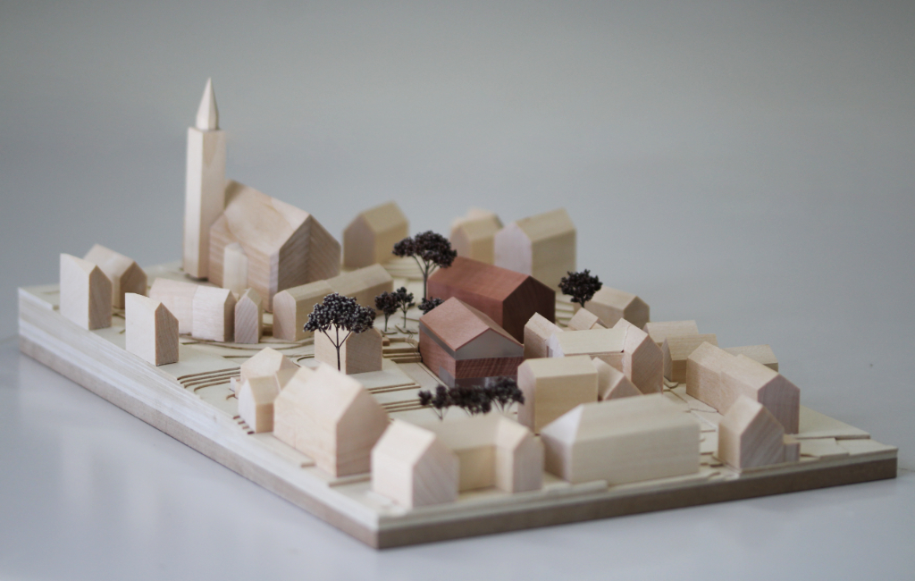 Modell für Architektenwettbewerb mit Höhenschichten aus gelblicher Finnpappe und Gebäuden aus Lindenholz. Der Neubau des Rathauses besteht aus zwei Gebäuden mit Satteldach und ist aus rötlichem Elsbeerenholz und mattiertem Acyl. Die Bäume sind aus dunkelbraunem getrocknetem Planzenmaterial.