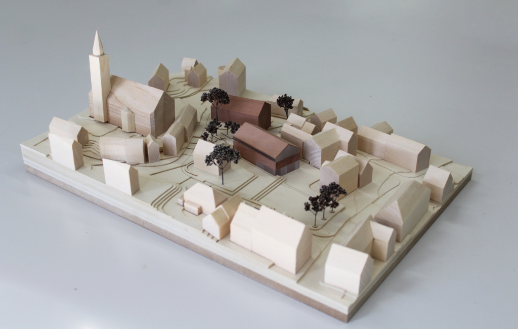 Modell für Architektenwettbewerb mit Höhenschichten aus gelblicher Finnpappe und Gebäuden aus Lindenholz. Der Neubau des Rathauses besteht aus zwei Gebäuden mit Satteldach und ist aus rötlichem Elsbeerenholz und mattiertem Acyl. Die Bäume sind aus dunkelbraunem getrocknetem Planzenmaterial.