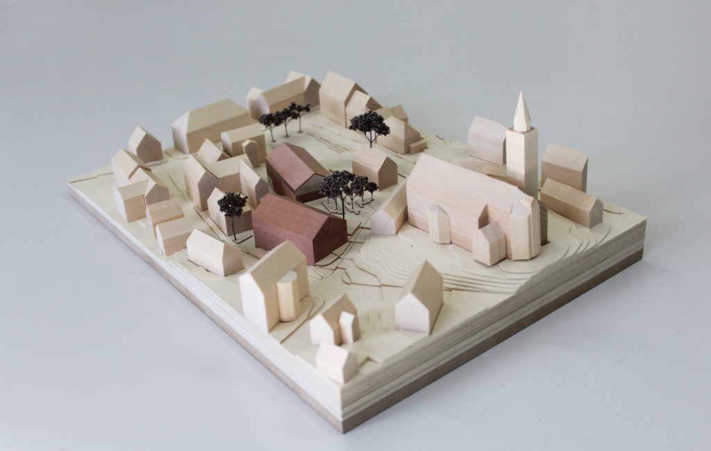 Modell für Architektenwettbewerb mit Höhenschichten aus gelblicher Finnpappe und Gebäuden aus Lindenholz. Der Neubau des Rathauses besteht aus zwei Gebäuden mit Satteldach und ist aus rötlichem Elsbeerenholz und mattiertem Acyl. Die Bäume sind aus dunkelbraunem getrocknetem Planzenmaterial.