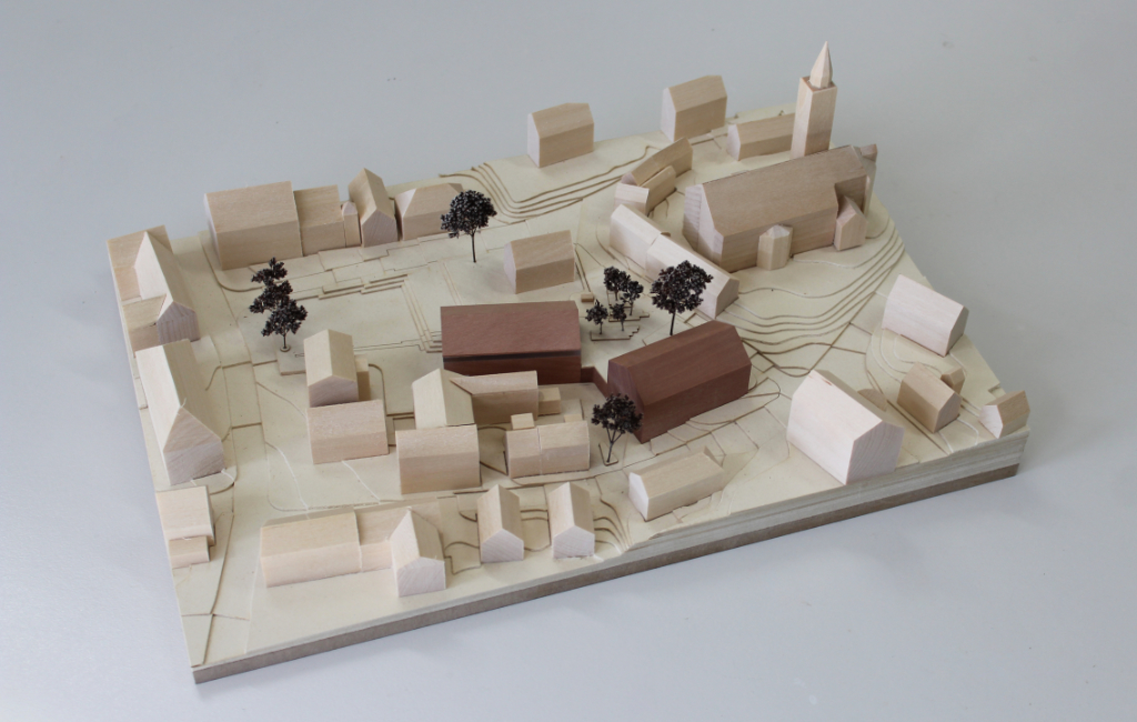 Modell für Architektenwettbewerb mit Höhenschichten aus gelblicher Finnpappe und Gebäuden aus Lindenholz. Der Neubau des Rathauses besteht aus zwei Gebäuden mit Satteldach und ist aus rötlichem Elsbeerenholz und mattiertem Acyl. Die Bäume sind aus dunkelbraunem getrocknetem Planzenmaterial.