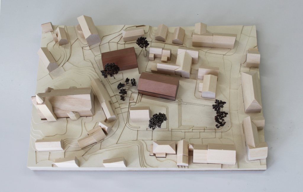 Modell für Architektenwettbewerb mit Höhenschichten aus gelblicher Finnpappe und Gebäuden aus Lindenholz. Der Neubau des Rathauses besteht aus zwei Gebäuden mit Satteldach und ist aus rötlichem Elsbeerenholz und mattiertem Acyl. Die Bäume sind aus dunkelbraunem getrocknetem Planzenmaterial.