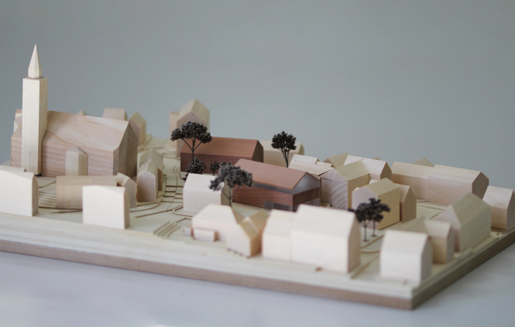 Modell für Architektenwettbewerb mit Höhenschichten aus gelblicher Finnpappe und Gebäuden aus Lindenholz. Der Neubau des Rathauses besteht aus zwei Gebäuden mit Satteldach und ist aus rötlichem Elsbeerenholz und mattiertem Acyl. Die Bäume sind aus dunkelbraunem getrocknetem Planzenmaterial.
