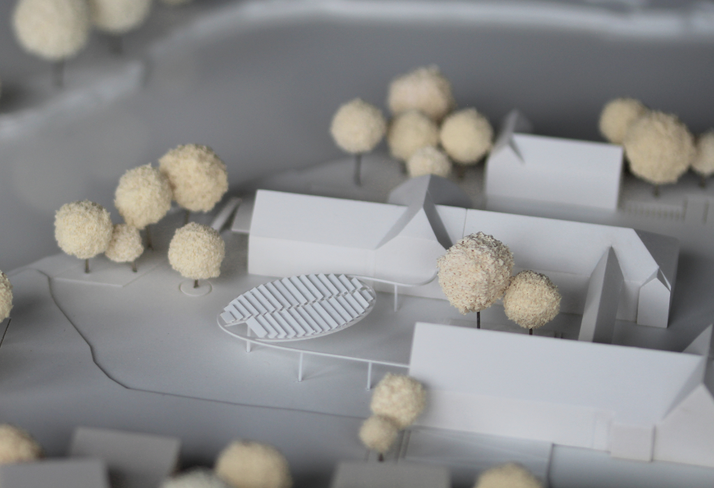 Fotografie von Modell für Architekturwettbewerb. Weißes Modell mit einfachen Gebäudekuben, Bäumen aus cremefarbenen Schwammgummikugeln, Wasser aus durchsichtigem Acrylglas, sowie einer detailliert dargestellten ellipsenförmigen Pergola mit Sonnenschutzelementen