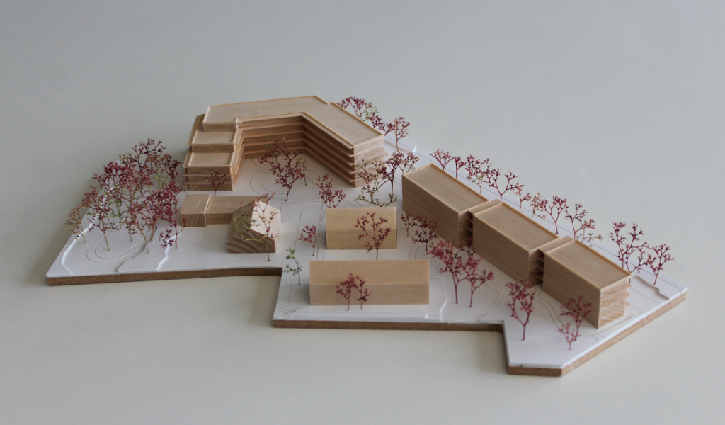 Modell für Architektenwettbewerb. Grundplatte aus weißem Kunststoff mit eingravierten Wegen. Gebäude aus massivem Lindenholz. Vier kleinere Gebäude mit Satteldach. Ein langgestreckter viergeschossiger Baukörper mit Flachdach entlang der Strasse. Ein hufeisenförmiger auf einer Seite abgetreppter fünfgeschossiger Baukörper mit Flachdach an der Spitze des Grundstücks. Bäume aus getrocknetem rötlichem Meerschaum.