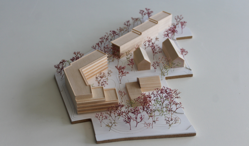 Modell für Architektenwettbewerb. Grundplatte aus weißem Kunststoff mit eingravierten Wegen. Gebäude aus massivem Lindenholz. Vier kleinere Gebäude mit Satteldach. Ein langgestreckter viergeschossiger Baukörper mit Flachdach entlang der Strasse. Ein hufeisenförmiger auf einer Seite abgetreppter fünfgeschossiger Baukörper mit Flachdach an der Spitze des Grundstücks. Bäume aus getrocknetem rötlichem Meerschaum.