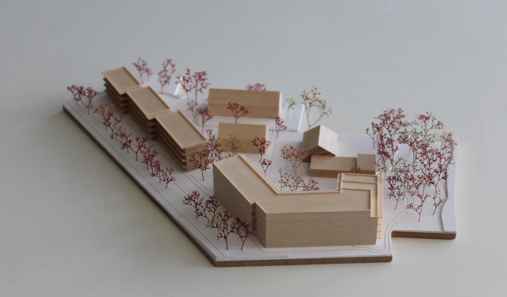 Modell für Architektenwettbewerb. Grundplatte aus weißem Kunststoff mit eingravierten Wegen. Gebäude aus massivem Lindenholz. Vier kleinere Gebäude mit Satteldach. Ein langgestreckter viergeschossiger Baukörper mit Flachdach entlang der Strasse. Ein hufeisenförmiger auf einer Seite abgetreppter fünfgeschossiger Baukörper mit Flachdach an der Spitze des Grundstücks. Bäume aus getrocknetem rötlichem Meerschaum.