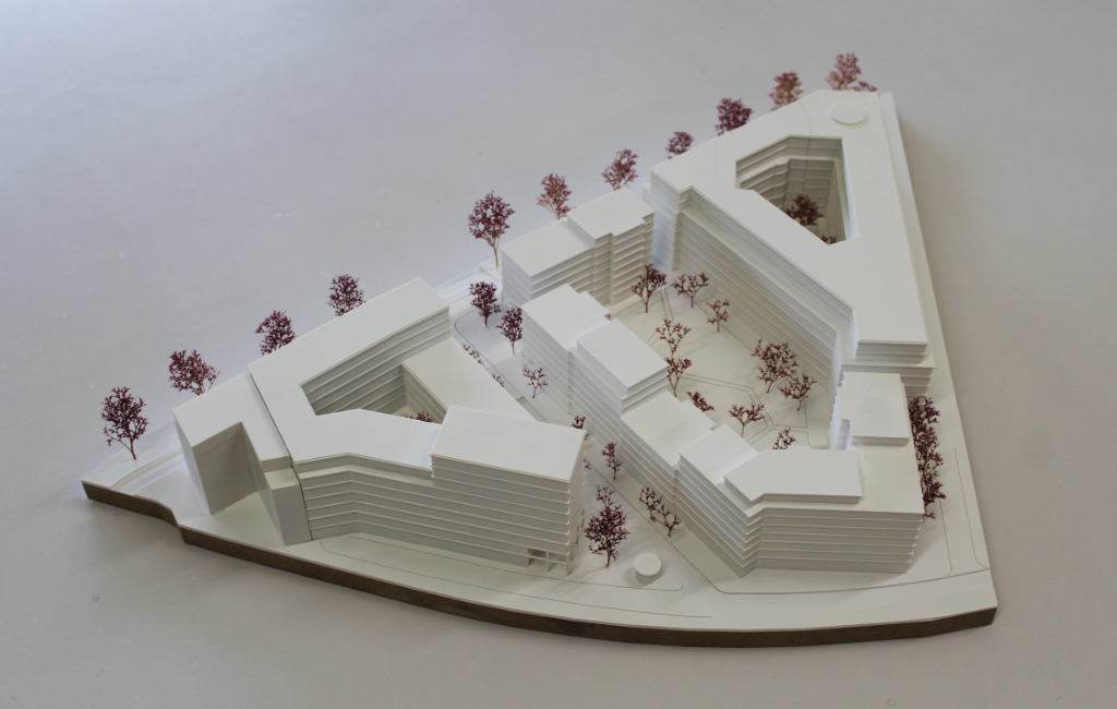 weißes architektonisches Massenmodell, bestehend aus vier großen Baukörpern angeordnet auf einer dreiecksförmigen Grundplatte. Die Geschosse der Baukörper sind ablesbar, da die Geschossdecken minimal auskragen. Die Bäume sind aus getrocknetem Pflanzenmaterial dargestellt. Das Modell ist von schräg oben fotografiert.