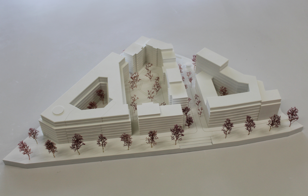 weißes architektonisches Massenmodell, bestehend aus vier großen Baukörpern angeordnet auf einer dreiecksförmigen Grundplatte. Die Geschosse der Baukörper sind ablesbar, da die Geschossdecken minimal auskragen. Die Bäume sind aus getrocknetem Pflanzenmaterial dargestellt. Das Modell ist von schräg oben fotografiert.