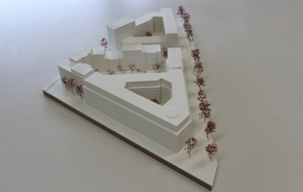 weißes architektonisches Massenmodell, bestehend aus vier großen Baukörpern angeordnet auf einer dreiecksförmigen Grundplatte. Die Geschosse der Baukörper sind ablesbar, da die Geschossdecken minimal auskragen. Die Bäume sind aus getrocknetem Pflanzenmaterial dargestellt. Das Modell ist von schräg oben fotografiert.