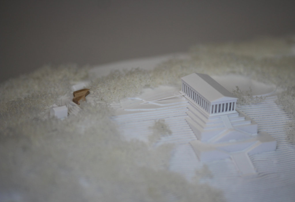 weißes Architekturmodell von der Walhalla und Umgebung. Der Höhenverlauf ist mittels Höhenschichten dargestellt, zur Dargestellung der Bäume ist weißer Pflanzenschaum flächig verklebt. Der Neubau des Besucherzentrums liegt nordöstlich von der Walhalla und besteht aus einem weißen Sockel auf dem ein kleines Holzvolumen mit mehreren Dachverschneidungen sitzt. Draufsicht von Süden.