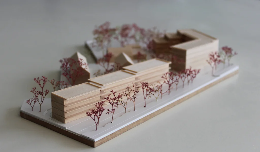 Modell für Architektenwettbewerb. Grundplatte aus weißem Kunststoff mit eingravierten Wegen. Gebäude aus massivem Lindenholz. Vier kleinere Gebäude mit Satteldach. Ein langgestreckter viergeschossiger Baukörper mit Flachdach entlang der Strasse. Ein hufeisenförmiger auf einer Seite abgetreppter fünfgeschossiger Baukörper mit Flachdach an der Spitze des Grundstücks. Bäume aus getrocknetem rötlichem Meerschaum.