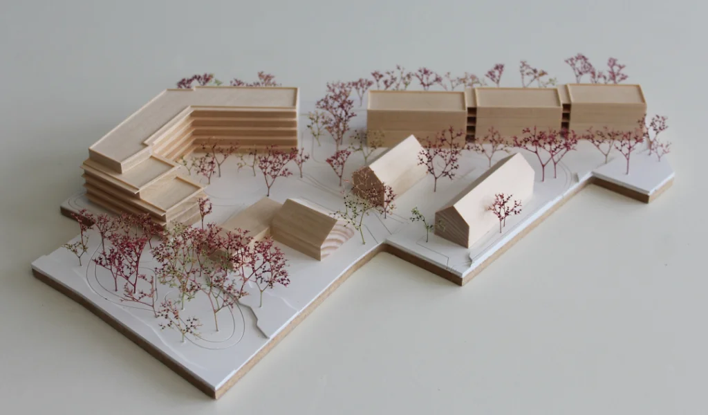 Modell für Architektenwettbewerb. Grundplatte aus weißem Kunststoff mit eingravierten Wegen. Gebäude aus massivem Lindenholz. Vier kleinere Gebäude mit Satteldach. Ein langgestreckter viergeschossiger Baukörper mit Flachdach entlang der Strasse. Ein hufeisenförmiger auf einer Seite abgetreppter fünfgeschossiger Baukörper mit Flachdach an der Spitze des Grundstücks. Bäume aus getrocknetem rötlichem Meerschaum.