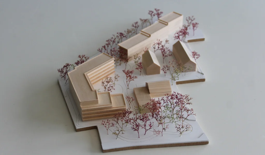 Modell für Architektenwettbewerb. Grundplatte aus weißem Kunststoff mit eingravierten Wegen. Gebäude aus massivem Lindenholz. Vier kleinere Gebäude mit Satteldach. Ein langgestreckter viergeschossiger Baukörper mit Flachdach entlang der Strasse. Ein hufeisenförmiger auf einer Seite abgetreppter fünfgeschossiger Baukörper mit Flachdach an der Spitze des Grundstücks. Bäume aus getrocknetem rötlichem Meerschaum.