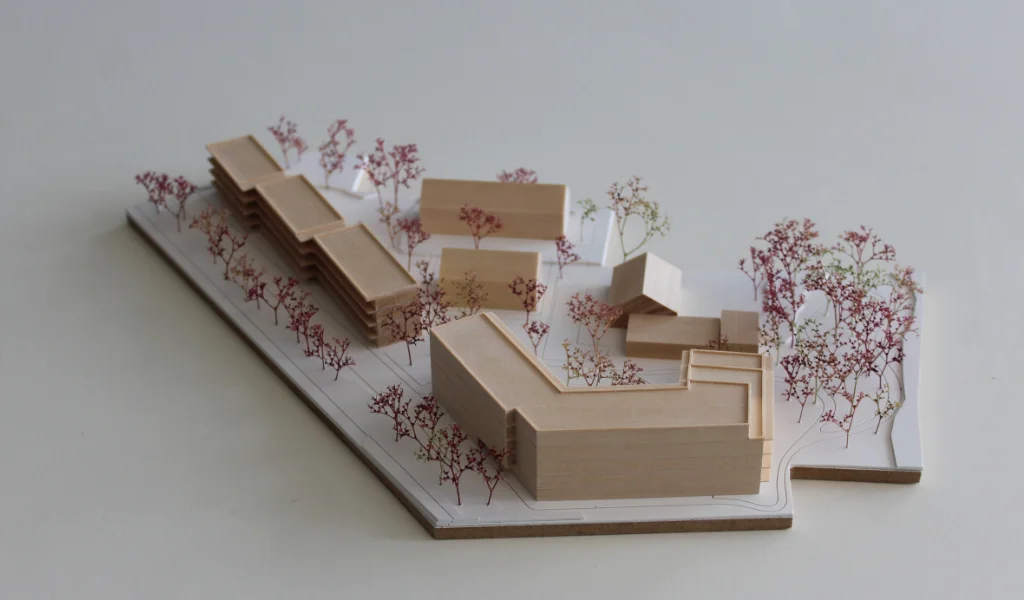 Modell für Architektenwettbewerb. Grundplatte aus weißem Kunststoff mit eingravierten Wegen. Gebäude aus massivem Lindenholz. Vier kleinere Gebäude mit Satteldach. Ein langgestreckter viergeschossiger Baukörper mit Flachdach entlang der Strasse. Ein hufeisenförmiger auf einer Seite abgetreppter fünfgeschossiger Baukörper mit Flachdach an der Spitze des Grundstücks. Bäume aus getrocknetem rötlichem Meerschaum.