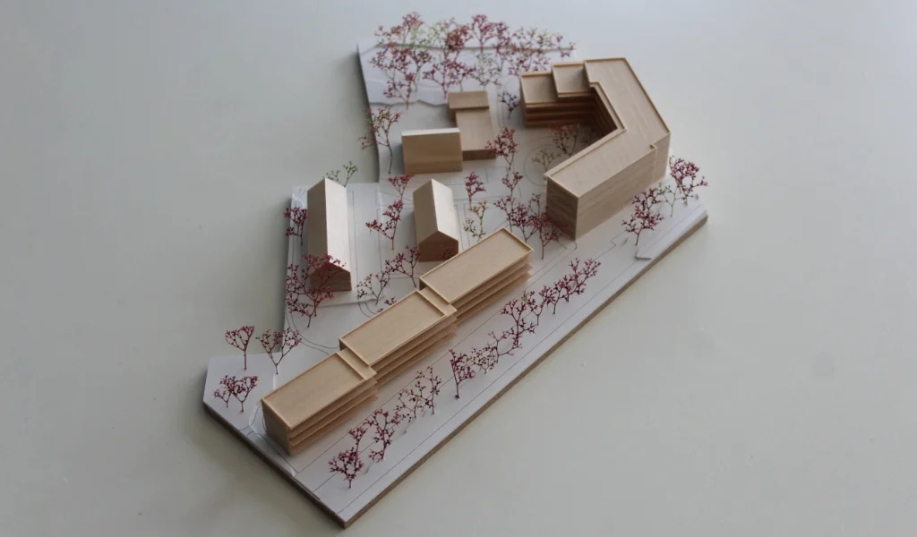 Modell für Architektenwettbewerb. Grundplatte aus weißem Kunststoff mit eingravierten Wegen. Gebäude aus massivem Lindenholz. Vier kleinere Gebäude mit Satteldach. Ein langgestreckter viergeschossiger Baukörper mit Flachdach entlang der Strasse. Ein hufeisenförmiger auf einer Seite abgetreppter fünfgeschossiger Baukörper mit Flachdach an der Spitze des Grundstücks. Bäume aus getrocknetem rötlichem Meerschaum.